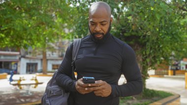 Yakışıklı, kel, sakallı Amerikalı adam güneşli bir günde şehir parkında telefon kullanıyor.