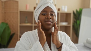 Genç, Afrikalı, Amerikalı, spa tedavisinden hoşlanan bir kadın, beyaz bir bornoz ve baş havlusu giyiyor, huzur veren bir sağlık merkezinde oturuyor..