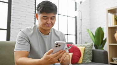 Genç Asyalı adam akıllı telefon kullanıyor ve modern apartman dairesinde kredi kartı tutuyor..