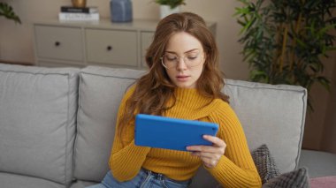 Genç, çekici, sarışın, beyaz, gözlüklü ve sarı kazaklı bir kadın gri bir kanepede otururken mavi bir tablet kullanıyor..