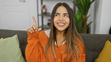 İçerideki gri koltukta oturan gülümseyen İspanyol kadın, parmağıyla yukarıya doğru işaret ediyor, turuncu bir kazak giyiyor..