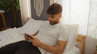 Young hispanic man with a beard lying on the bed in a bedroom holding a smartphone indoors