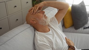 Oturma odasının iç tarafındaki koltukta dinlenen gri saçlı ve sakallı olgun bir adam. Alnında bir elle bitkin görünüyor.