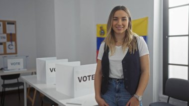 Güzel, genç, sarışın bir kadın Venezuela 'da bir seçim salonunda oylama düzeniyle duruyor. Arka planda Venezuela bayrağı olan kapalı bir oylama ortamı sergiliyor..