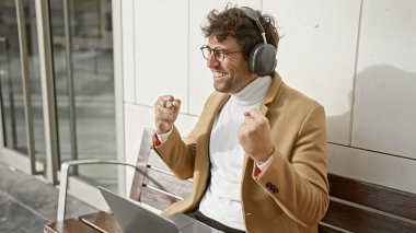 Kulaklıklı gülümseyen adam, şehir bankında dizüstü bilgisayarla başarıyı kutluyor..