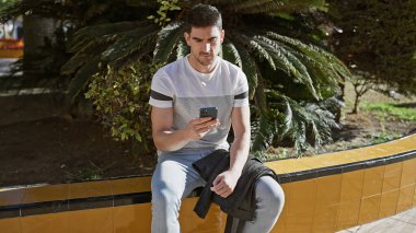 Yakışıklı İspanyol adam güneşli bir parkta bankta otururken akıllı telefon kullanıyor..