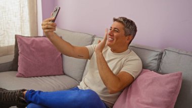 Genç bir adam oturma odasında video konferansı yapıyor, pembe ve gri yastıklarla bir koltukta oturuyor, akıllı telefonuna el sallıyor ve gülümsüyor..
