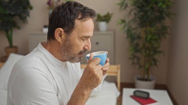 Orta yaşlı İspanyol bir adam rahat bir yatak odasında kahve içiyor. Çiçekler ve sabah ışığı içeri giriyor..