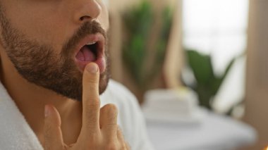 Kapalı bir güzellik merkezinde sakallı genç İspanyol bir adam parmaklarıyla dudağına dokunuyor.