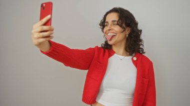 Genç, güzel, İspanyol bir kadın selfie çekiyor ve beyaz bir arka planda şakacı bir şekilde poz verirken dilini dışarı çıkarıyor..