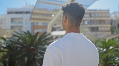 Yakışıklı İspanyol genç adam güneşli bir şehir parkında duruyor, modern bir şehir caddesine ve canlı bir kasabanın dışındaki palmiye ağaçlarına bakıyor..