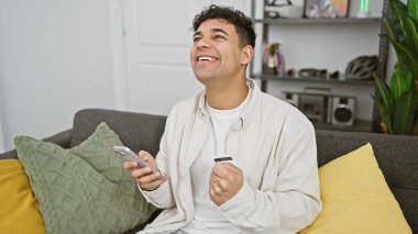 Genç bir adam, elinde bir kredi kartı ve akıllı telefon tutarak oturma odasından başarılı bir online alışveriş kutluyor..