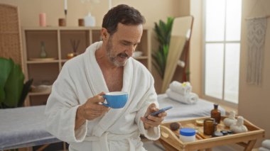 Sağlıklı spa odasında rahatlayan, bornoz giyen ve akıllı telefon kullanırken mavi bardaktan içen olgun İspanyol bir adam.