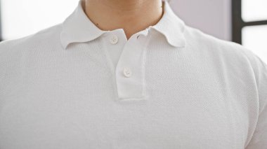 Parlak bir odada beyaz polo tişörtü giyen genç bir adam, sıradan ve düzenli bir görünüm sergiliyor..