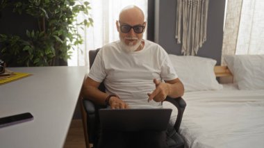 Olgun bir adam evinde bir yatak odasında videollamada, kucağında dizüstü bilgisayarla bir sandalyede oturuyor, gözlük takıyor ve beyaz bir gömlek giyiyor, baş parmağını kaldırıyor..