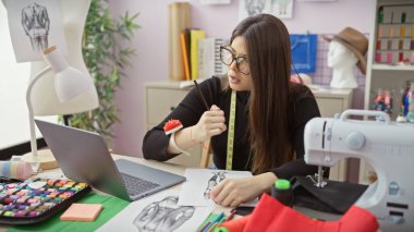 Genç bir kadın atölyesinde el işi yaparken ölçü almaya odaklanır. Moda çizimleri ve dikiş makinesiyle çevrili..