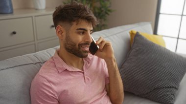 Oturma odasındaki kanepede otururken telefonda konuşan sakallı İspanyol bir genç..