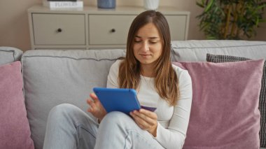 Sarışın, genç, çekici bir kadın oturma odasında rastgele oturuyor, elinde kredi kartı tutarken tablet kullanıyor, dairesindeki rahat bir koltukta minderlerle çevrili..