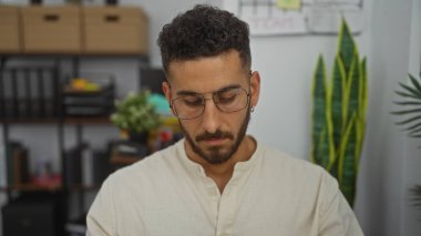 Yakışıklı, gözlüklü, iç mekan bitkileriyle dolu bir ofiste oturan ve odaklanmış bir ifade takınan bir İspanyol..