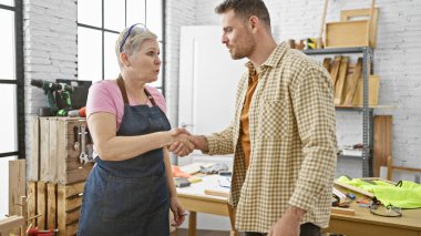 Bir adam ve bir kadın ahşap atölyesinde el sıkışıyor. Etrafı alet ve kerestelerle çevrili..