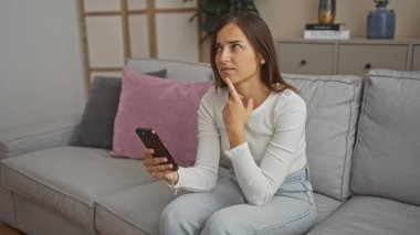 Oturma odasında oturan genç, güzel bir kadın, elinde akıllı bir telefonla düşünceli görünüyor, rahat, modern bir daire ile çevrili..