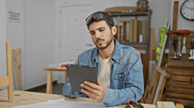 Sakallı genç bir İspanyol, iyi donanımlı bir marangoz atölyesinde düşünceli bir şekilde tablet kullanıyor..