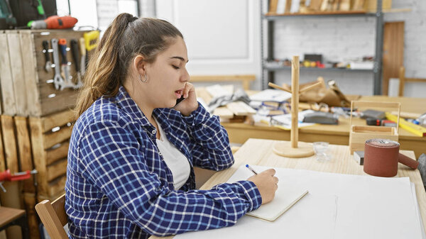Задумчивая молодая женщина рисует проекты в хорошо оборудованной столярной мастерской с видимыми инструментами и деревом.
