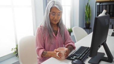 Ofiste çalışan, elinde kahve fincanı olan, gri saçlı, masasında oturan, etrafı bilgisayar, bitki ve raflarla çevrili yaşlı bir kadın..