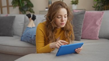 Genç ve çekici sarışın bir kadın evdeki bir evin oturma odasındaki kanepede dinlenirken tablet kullanıyor..