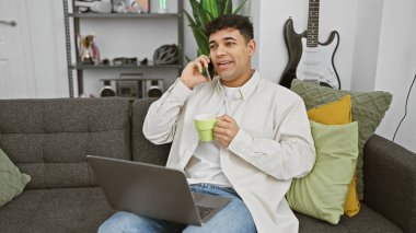 Sıradan bir adam elinde bir kupa, dizüstü bilgisayarı açık ve arka planda rahat bir oturma odasında gitar tutarken telefonla konuşur..