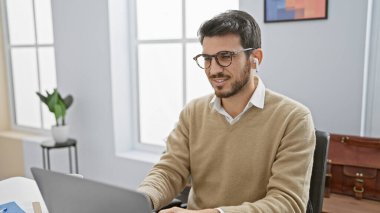 İspanyol adam gülümsüyor, modern bir ofiste dizüstü bilgisayar kullanıyor, profesyonel, stil sahibi ve üretken bir atmosferi canlandırıyor..