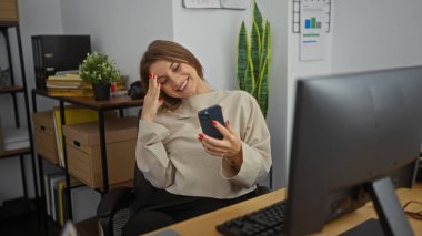 Genç, güzel, sarışın, beyaz bir kadın telefonuna bakarken gülümsüyor modern ofis ortamında raflar, bitkiler ve ofis malzemeleriyle..