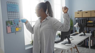 Siyah bir kadın parlak bir ofiste elinde çalışma masası olan mavi bir kahve fincanı ve duvarda fikir tahtasıyla geriniyor.