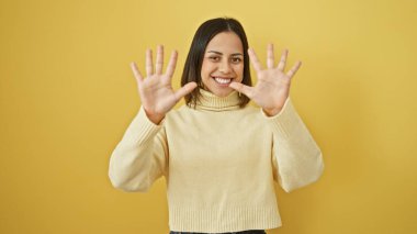 Krem rengi kazaklı genç bir kadın gülümseyerek poz veriyor ve sarı arka planda 10 numarayı işaret ediyor..