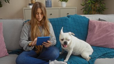 Genç sarışın bir kadın oturma odasında köpeğiyle oturuyor, renkli yastıklarla kanepede otururken tablet kullanıyor..
