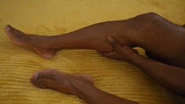 Yatak odasında oturan Afrikalı bir Amerikalı evindeki sarı yatakta otururken bacağını tutuyor.