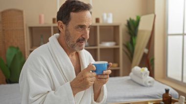 Beyaz bornozlu olgun İspanyol bir adam. Huzurlu bir spa odasında bir fincan kahvenin keyfini çıkarıyor. Terapi amaçlı dekor ve sağIıklı bir ortam..