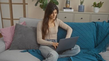 Genç İspanyol esmer kadın rahat bir oturma odasında dizüstü bilgisayarla çalışıyor. Modern dekorları ve çiçekleri var. Renkli yastıkları olan rahat bir koltukta oturuyor..