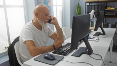 Ofiste bilgisayarda çalışan, kel, gri saçlı ve sakallı bir adam, telefonda konuşurken evde oturuyor, modern işyerinde pencereleri, bitkileri ve ofis malzemeleri ile gülümsüyor.