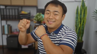 Yakışıklı genç Asyalı bir adam arka planda bitki ve raflarla bir ofis odasında güvenle gülümsüyor..