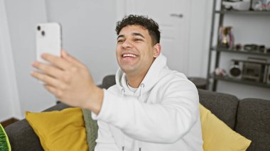 Gülümseyen genç bir adam akıllı telefonuyla kapalı bir salonda beyaz kapüşonlu bir ceket giyerek selfie çekiyor.