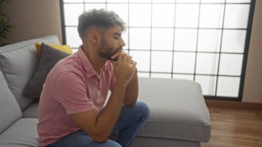 Yakışıklı, sakallı genç bir adam. Oturma odasında oturmuş, elleri çenesinde düşünceli bir şekilde düşünüyor.