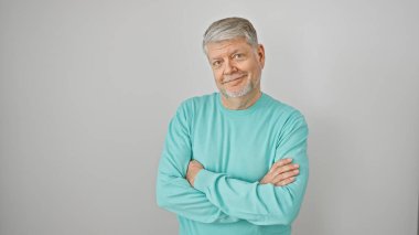 Gri saçlı ve turkuaz süveterli orta yaşlı bir adam kollarını beyaz bir arka plana çarpmış, kendine güveni ve olgunluğu yayıyor..