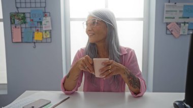 Olgun, gri saçlı bir kadın ofis ortamında elinde bardakla gülümsüyor. Etrafı notlar ve kağıtlarla çevrili. İş ortamında verimlilik ve rahatlığı resmediyor..