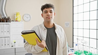 Handsome young man with a beard holding a clipboard in a bright laboratory setting. clipart