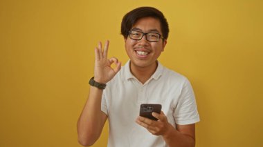 Genç Çinli adam gülümsüyor ve iyi bir hareket sergiliyor. Elinde akıllı telefon tutarken, sarı arka planda izole edilmiş.