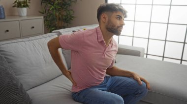 Young man with a beard in a pink shirt sitting in a living room and holding his back in pain, suggesting discomfort or injury in an indoor home setting clipart