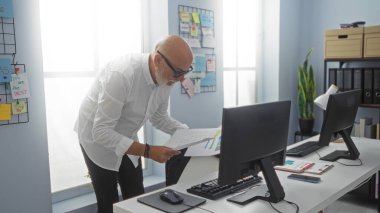 Gri saçlı ve sakallı orta yaşlı bir adam modern bir ofiste çalışıyor, elinde belgelerle bilgisayar ekranının önünde duruyor..