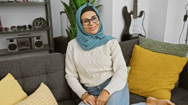 A smiling mature woman wearing a hijab sitting comfortably in a modern living room on a gray couch with colorful pillows. clipart