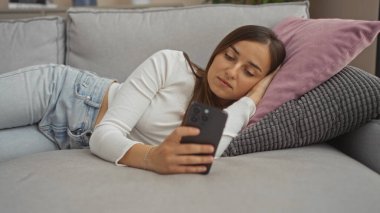 Evde rahat rahat oturan, akıllı telefon kullanarak kanepede uzanan genç bir kadın..